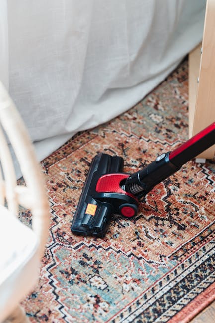 Close-up view of a vacuum cleaner head positioned on a patterned area rug with floral motifs, situated in a well-lit room. The carpet appears clean and well-maintained, with a person’s legs visible