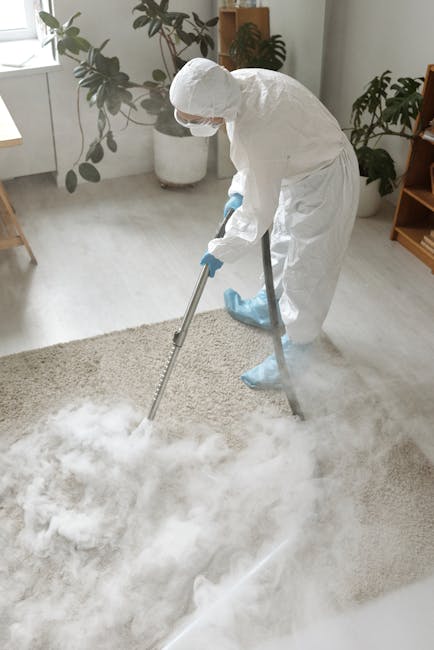 A professional cleaner dressed in a white protective suit, gloves, and shoe covers is performing surface cleaning with a vacuum or extraction device on a beige carpet in a well-lit, modern living room