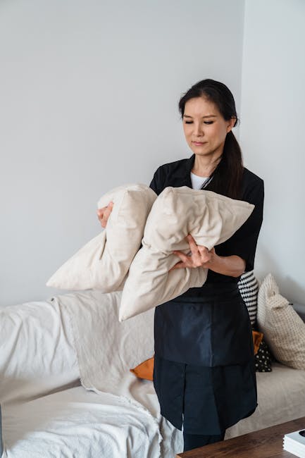 A person wearing black gloves is using a handheld electric sander on a dark grey fabric sofa, which is situated in a well-lit living room. The sofa has a modern design with a cushioned armrest on the